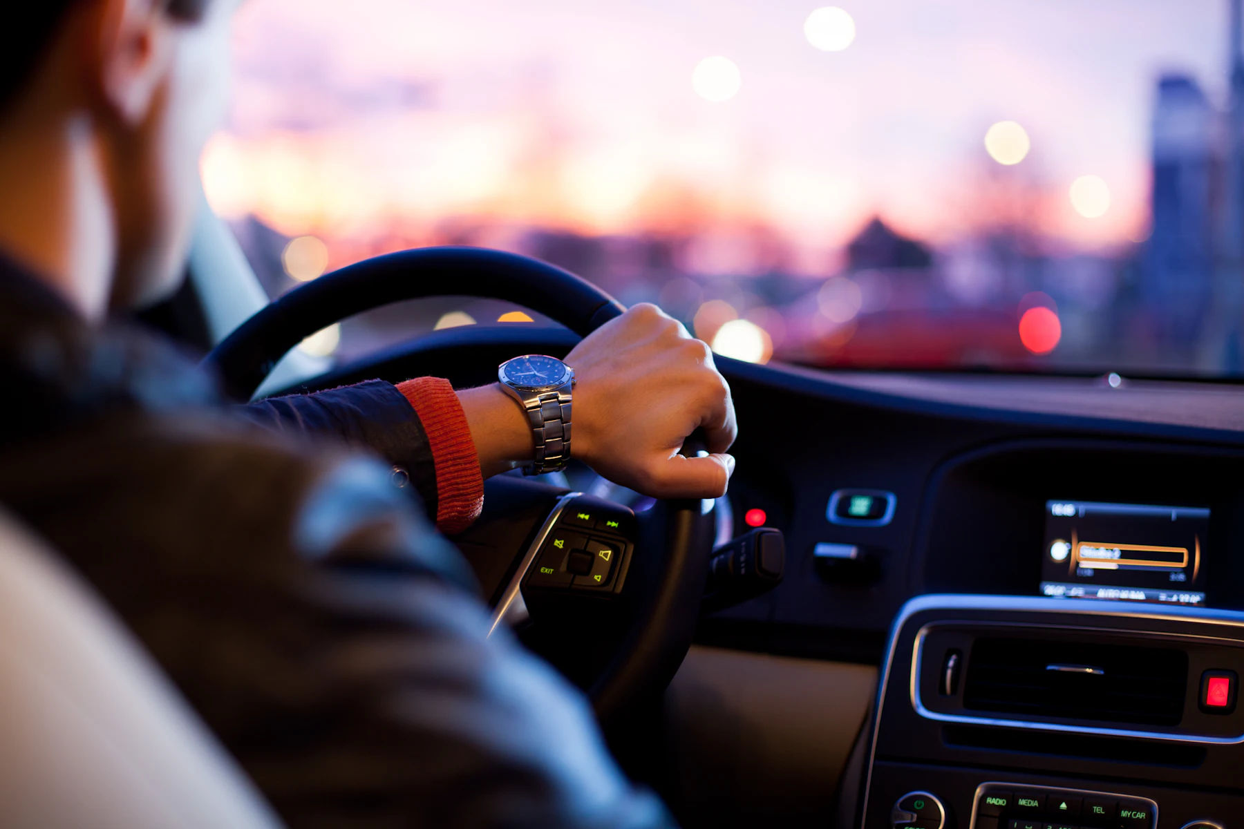 A driver's hands on the steering wheel at dusk, city lights blurred ahead.