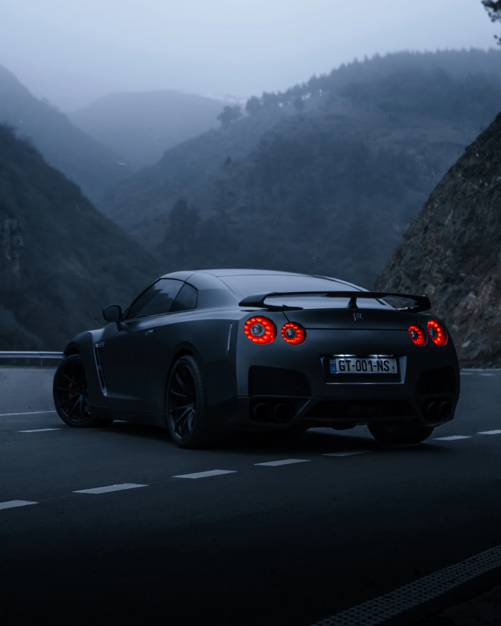 A dark sports car with glowing round tail lights on a mountain road at dusk.
