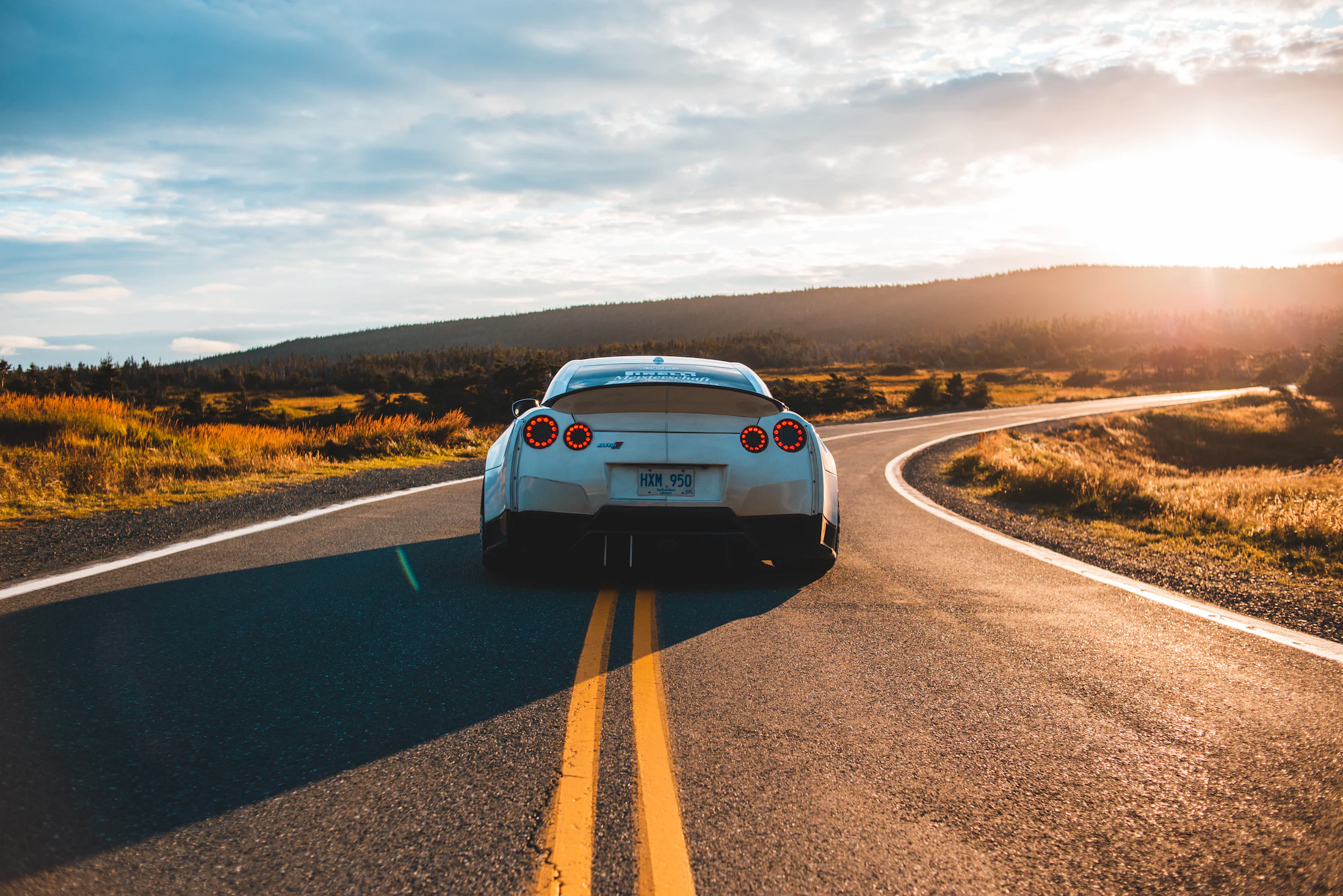 A silver sports car on a winding two-lane road toward a low sun.
