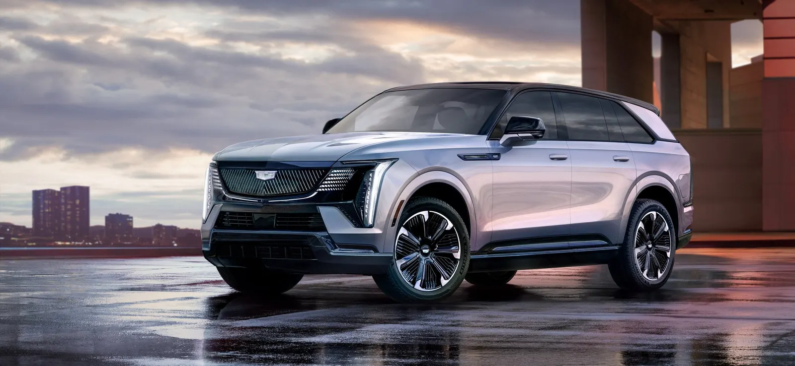 A silver Cadillac Escalade IQ electric SUV on wet pavement with a city skyline at dusk behind it