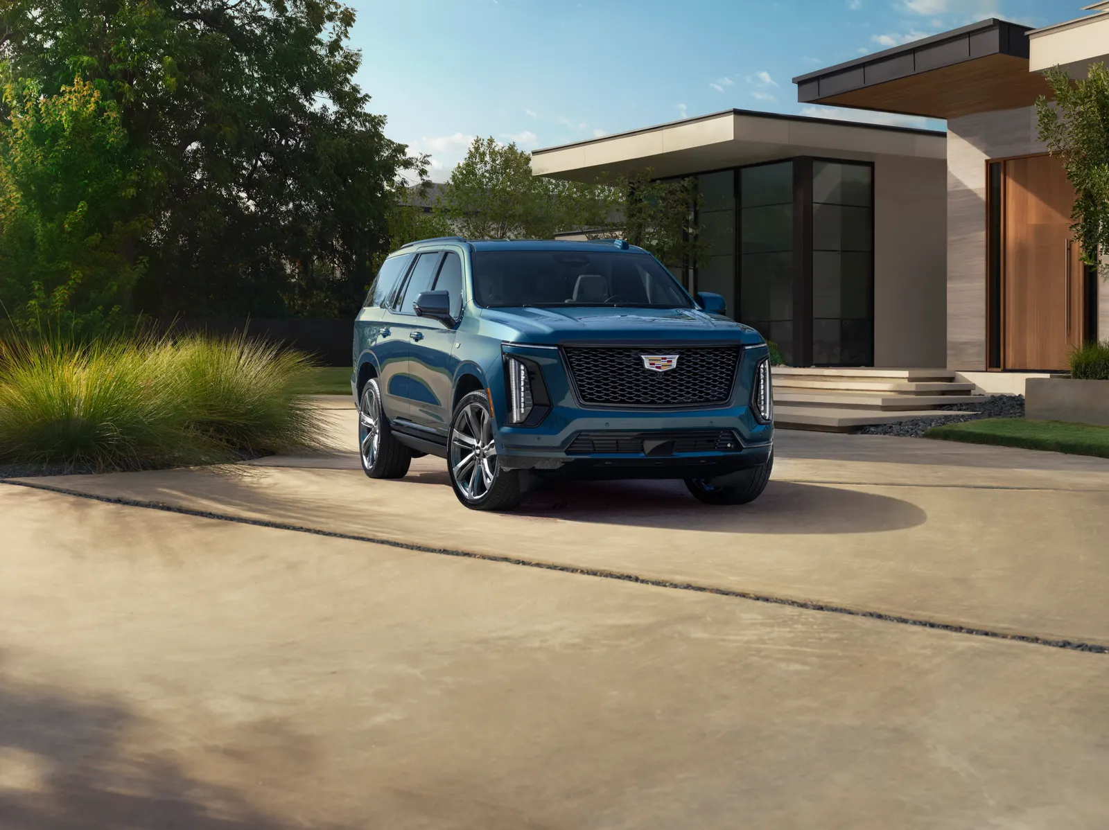A blue Cadillac Escalade with the updated 2025 front end parked on a concrete driveway outside a modern house