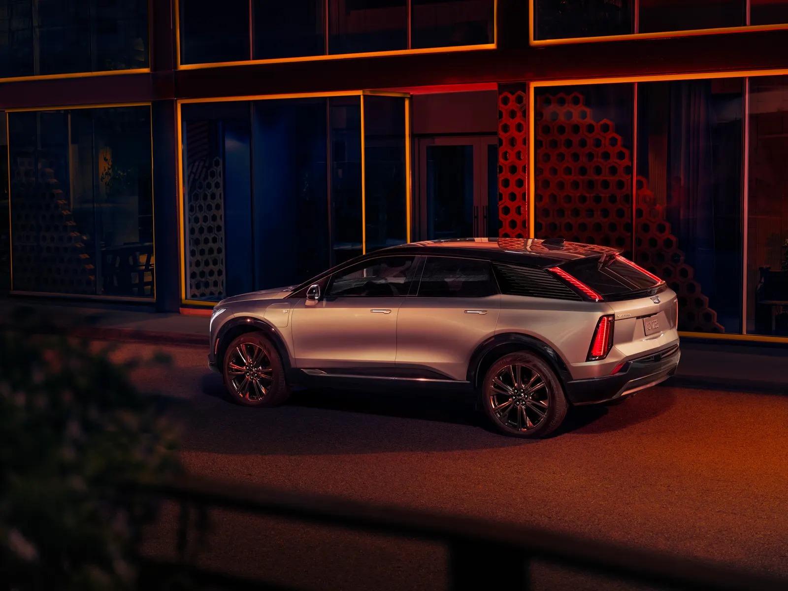 Rear three-quarter view of a silver Cadillac Optiq parked on a street at night beside a building with lit orange windows