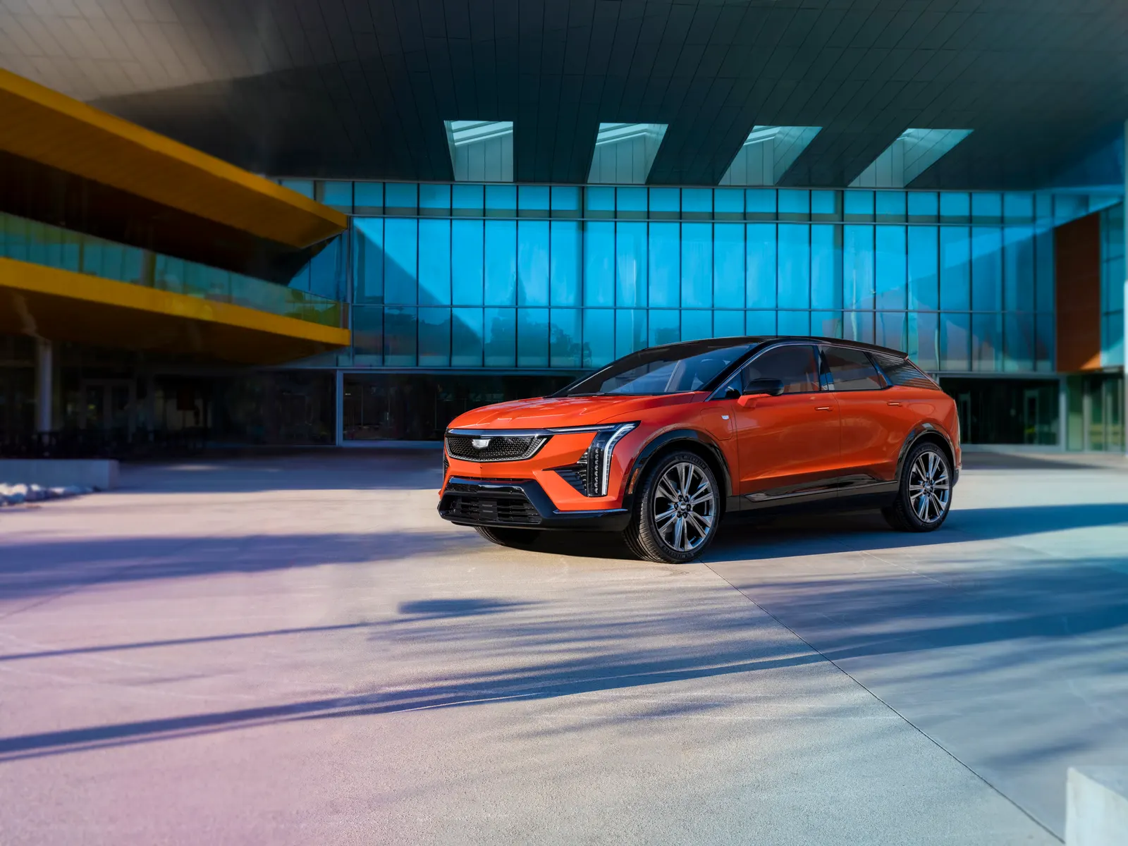 An orange Cadillac Optiq electric SUV parked on a sunlit plaza in front of a glass-walled building