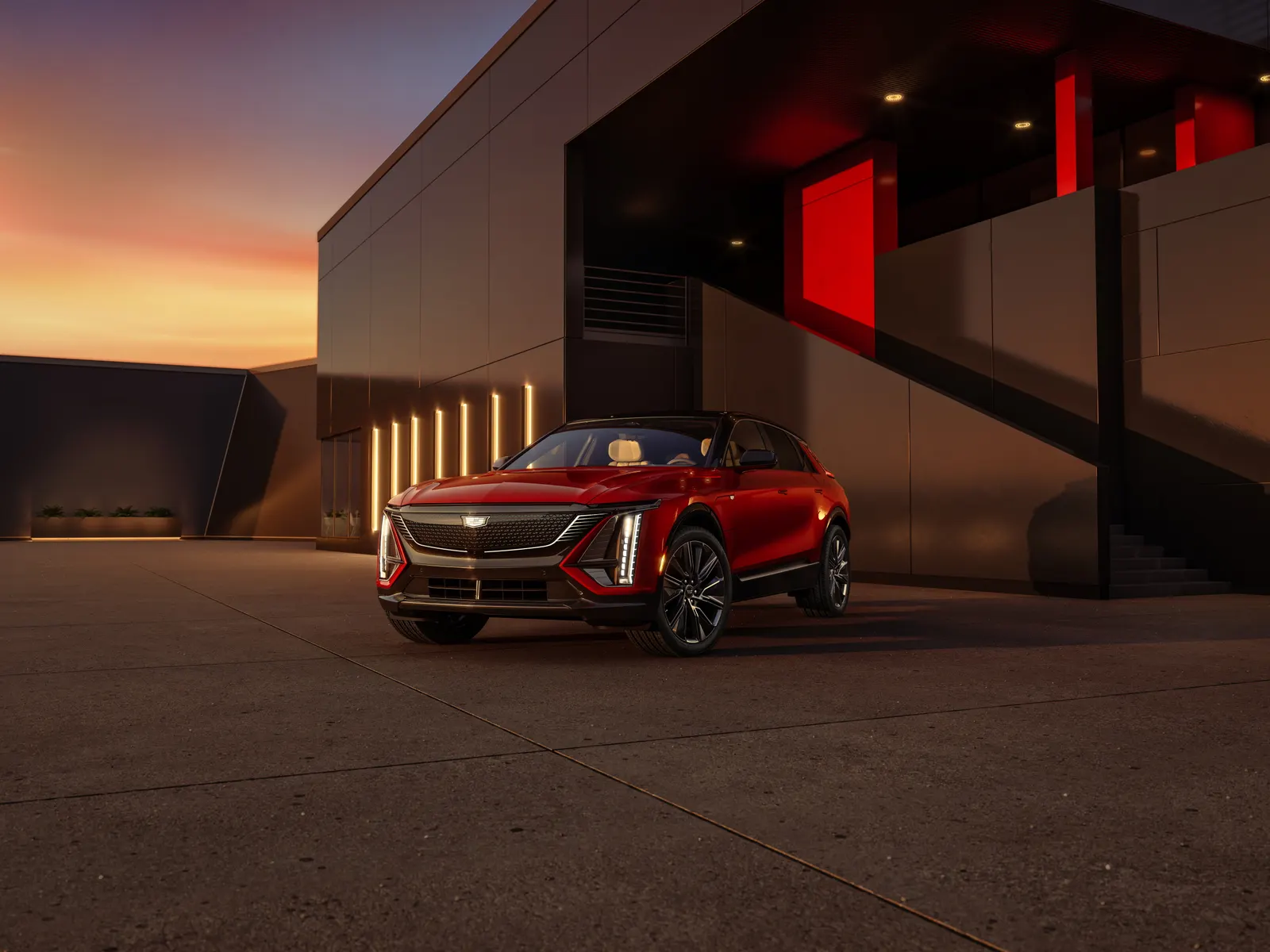 A red 2024 Cadillac Lyriq parked at dusk in front of a modern building with lit vertical panels