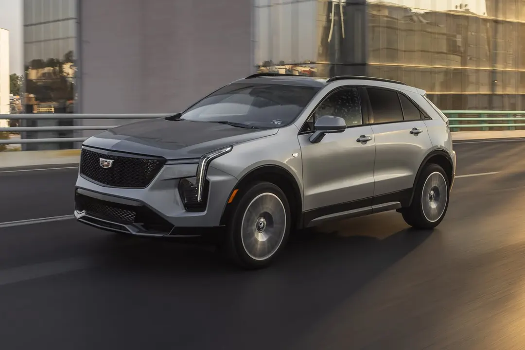 A silver 2024 Cadillac XT4 Sport with a black roof driving across an overpass in front of glass buildings
