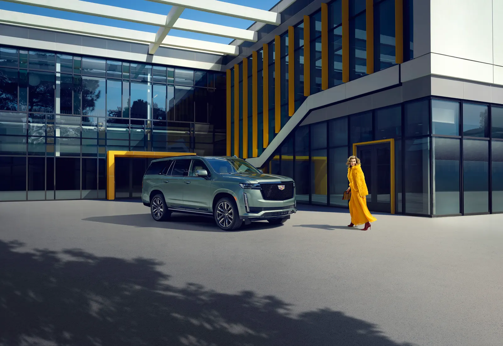 A gray-green 2024 Cadillac Escalade parked outside a glass building as a woman in a yellow coat walks past