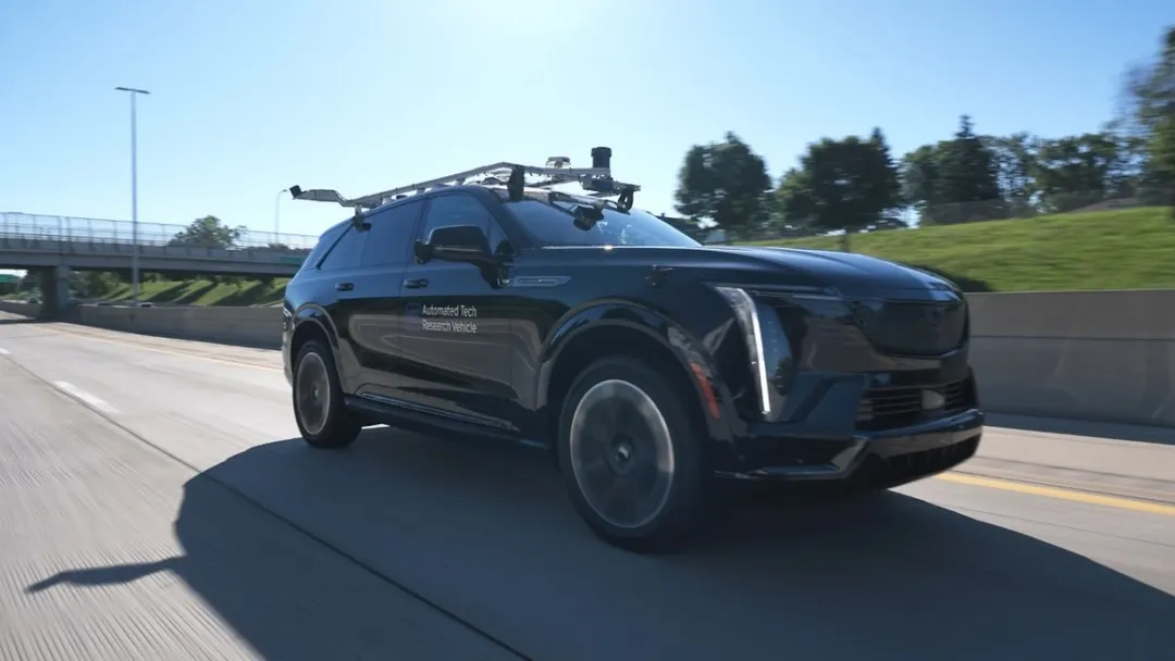 A black Cadillac Escalade IQ marked Automated Tech Research Vehicle drives on a highway with a sensor rack on its roof