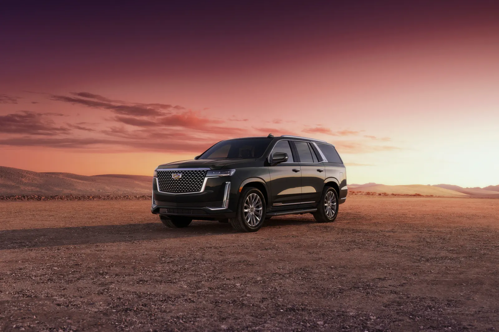 A black Cadillac Escalade of the 2021-2024 design parked on a desert plain under a pink and purple sunset sky