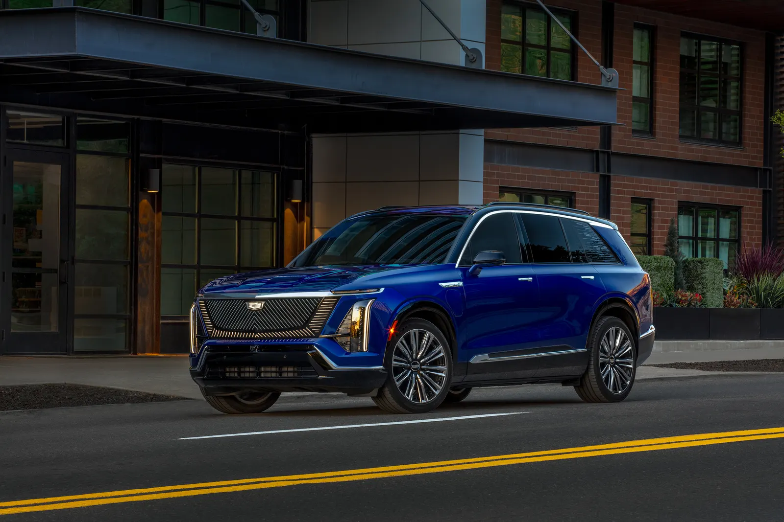 A blue Cadillac Vistiq electric SUV driving on a city street past brick buildings at dusk