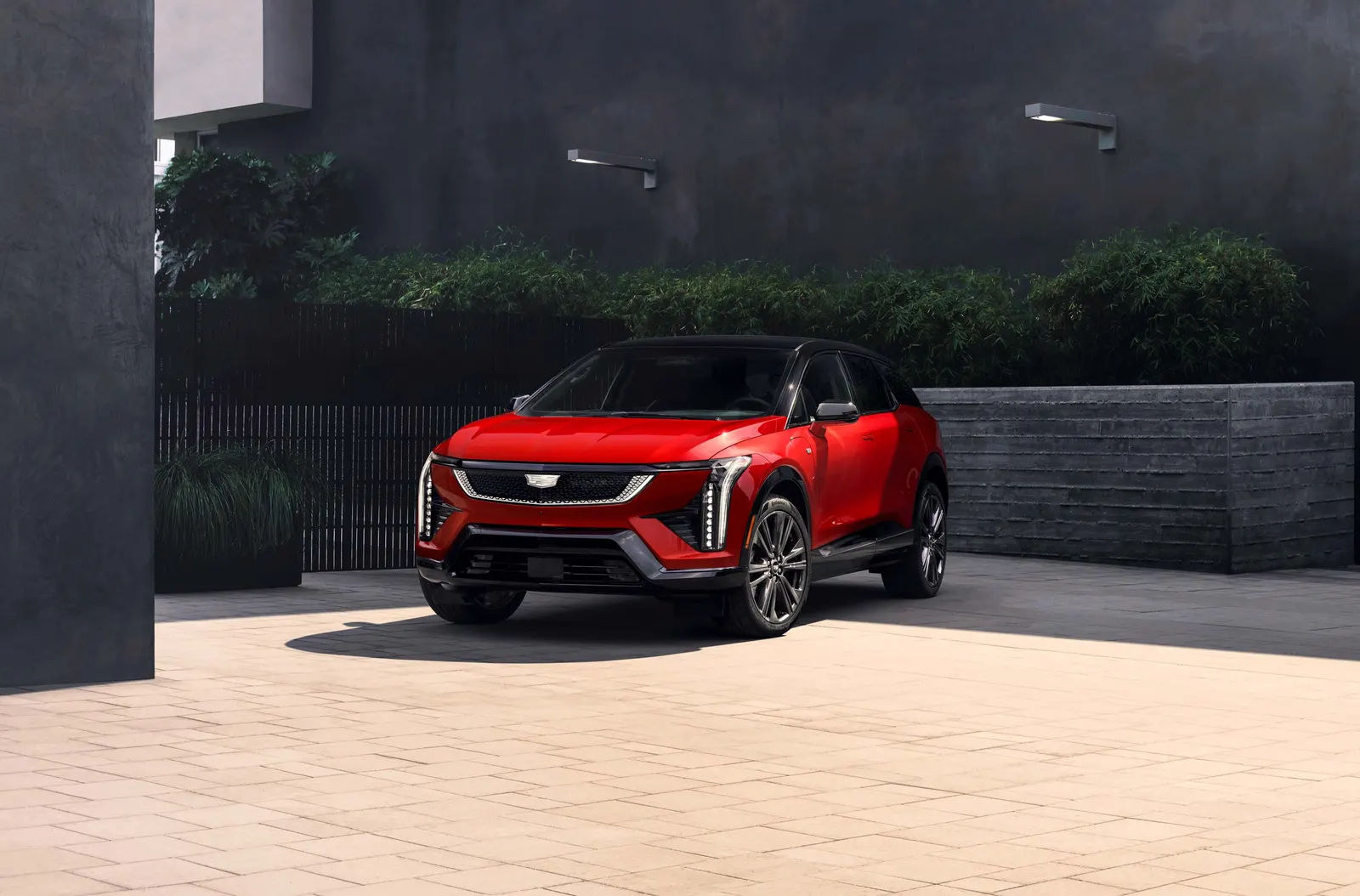 Red 2027 Cadillac Optiq with a black roof parked on a pale stone courtyard beside dark walls and hedges