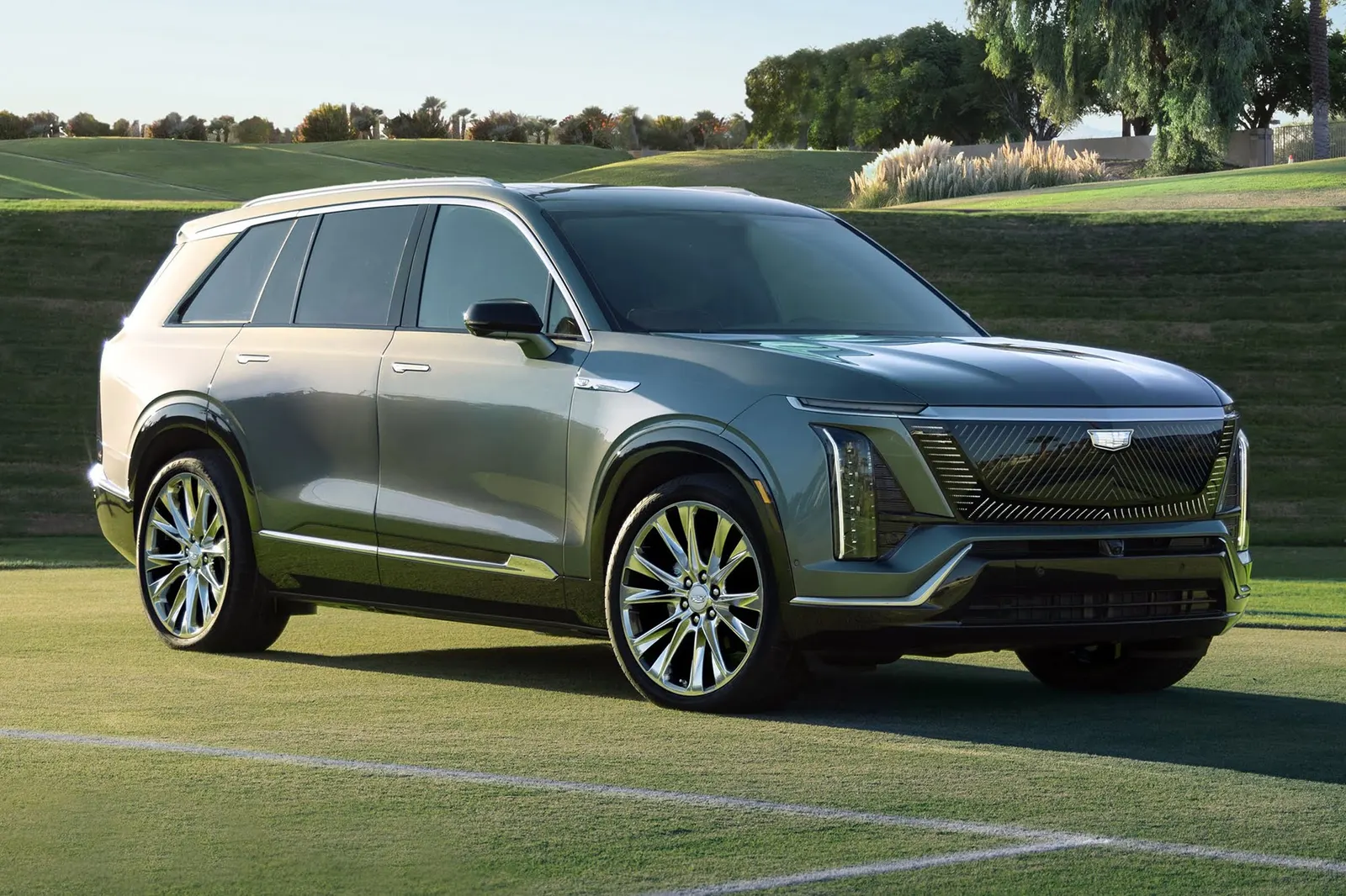 A gray-green Cadillac Vistiq three-row electric SUV with chrome wheels parked on a grass field, seen from the front three-quarter angle