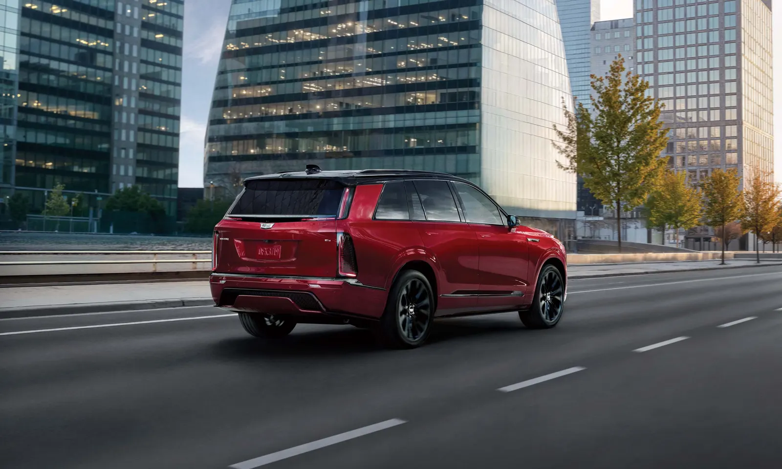 Rear three-quarter view of a red 2027 Cadillac Vistiq driving on a multi-lane city road past glass office towers