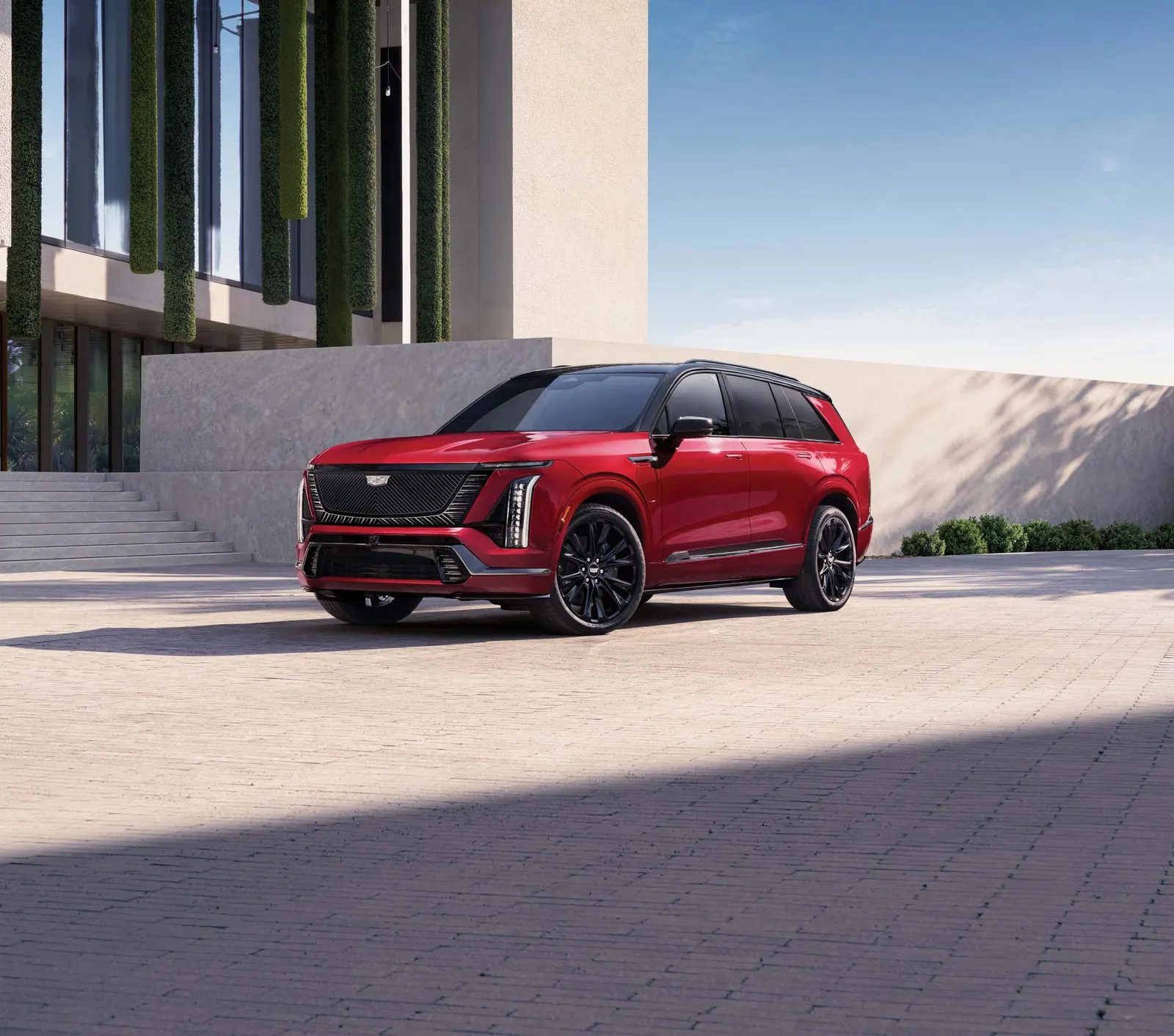 A red 2027 Cadillac Vistiq with black wheels parked on a brick plaza in front of a modern building with hanging greenery