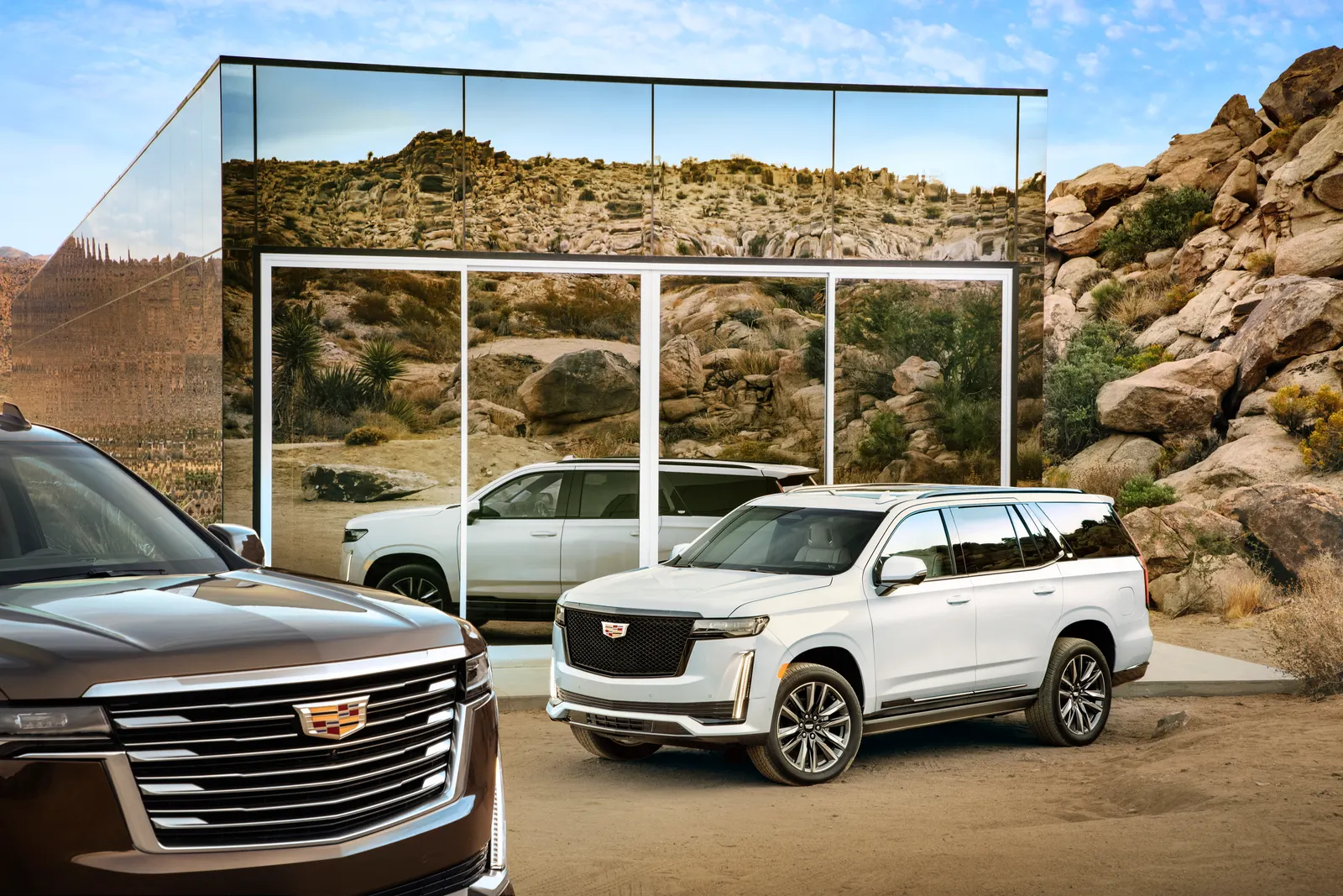 A white Cadillac Escalade parked beside a mirrored glass building among desert boulders, with a dark Escalade in the foreground