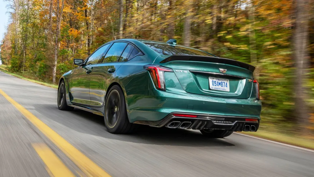 A green 2026 Cadillac CT5-V Blackwing sedan driving on a two-lane road lined with autumn trees, seen from the rear