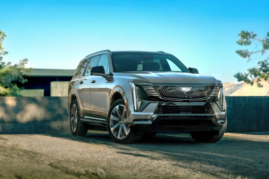 A silver Cadillac Escalade IQL electric SUV parked on gravel in front of a low wall under a clear blue sky