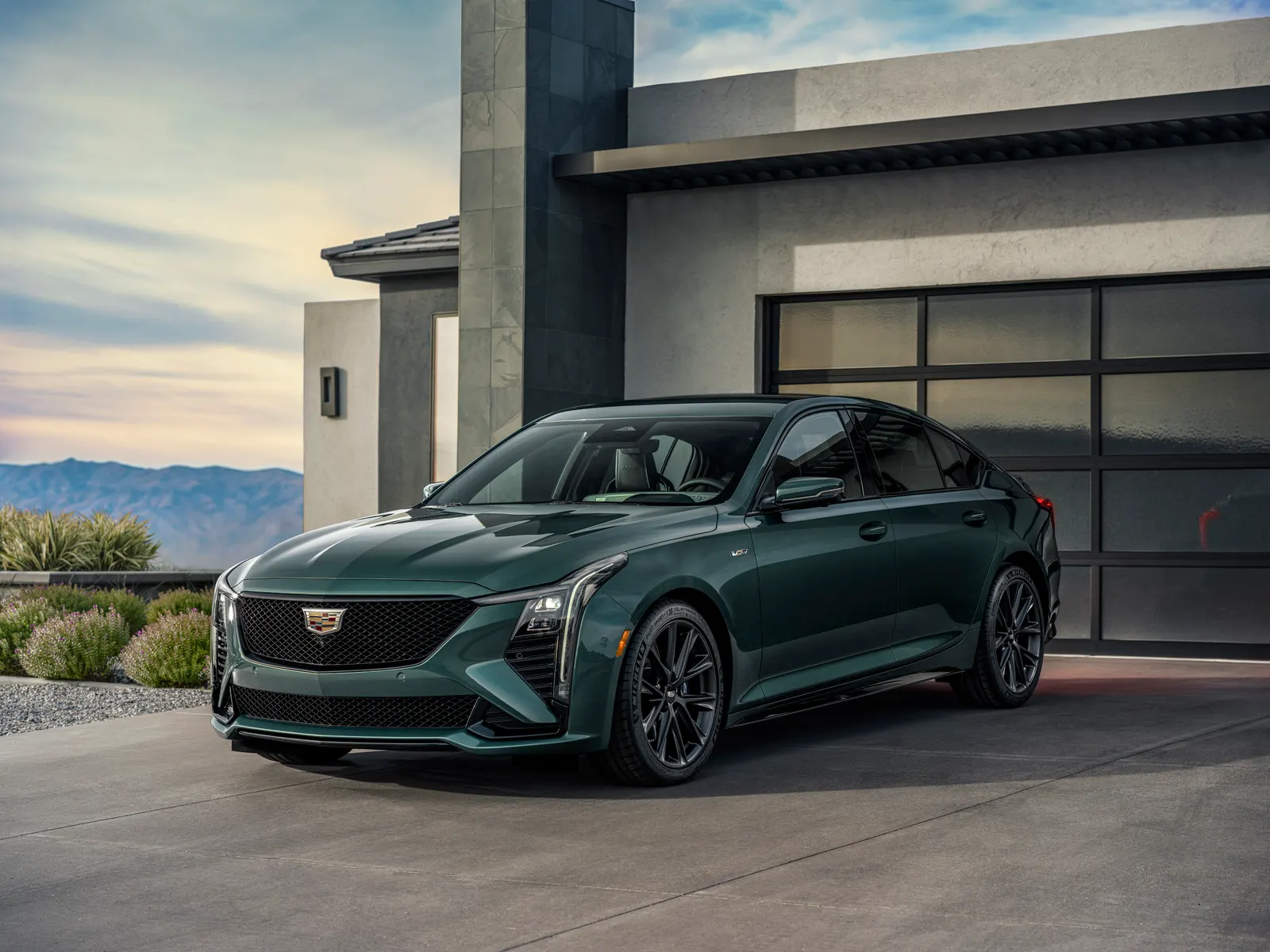A dark green Cadillac CT5-V sedan with black wheels parked in front of a modern house and garage at dusk