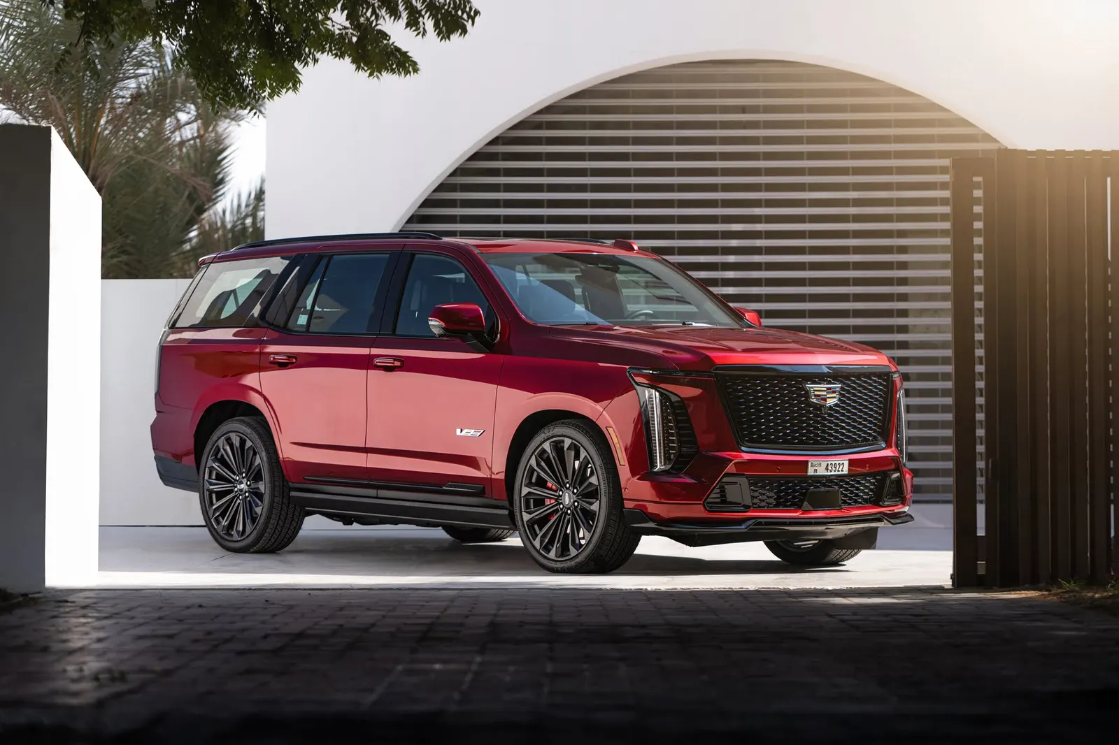 A red 2025 Cadillac Escalade-V on large dark multi-spoke wheels, parked under an arched white wall