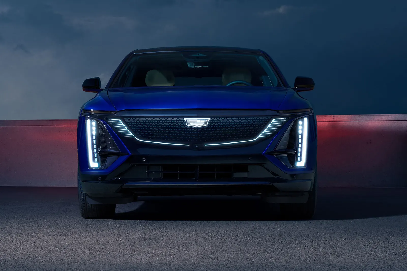 A blue Cadillac Lyriq electric SUV seen head-on at dusk with its vertical LED headlights lit