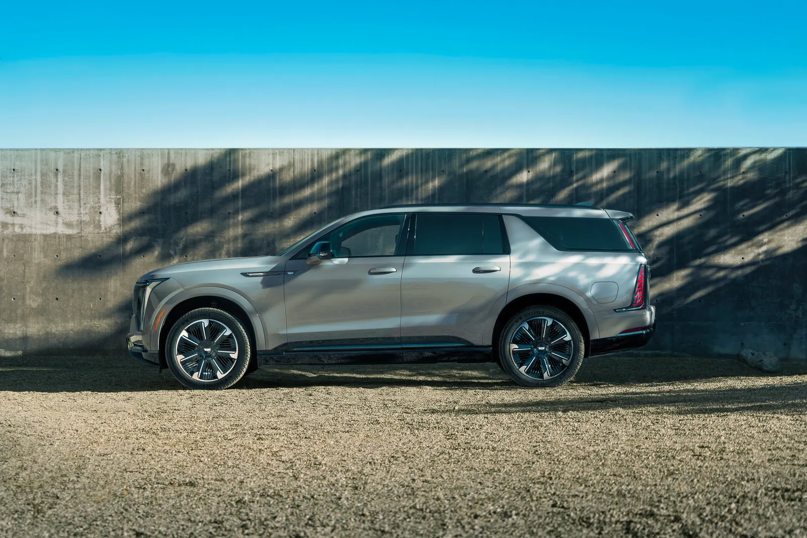 A silver 2026 Cadillac Escalade IQL parked in side profile in front of a concrete wall under a blue sky