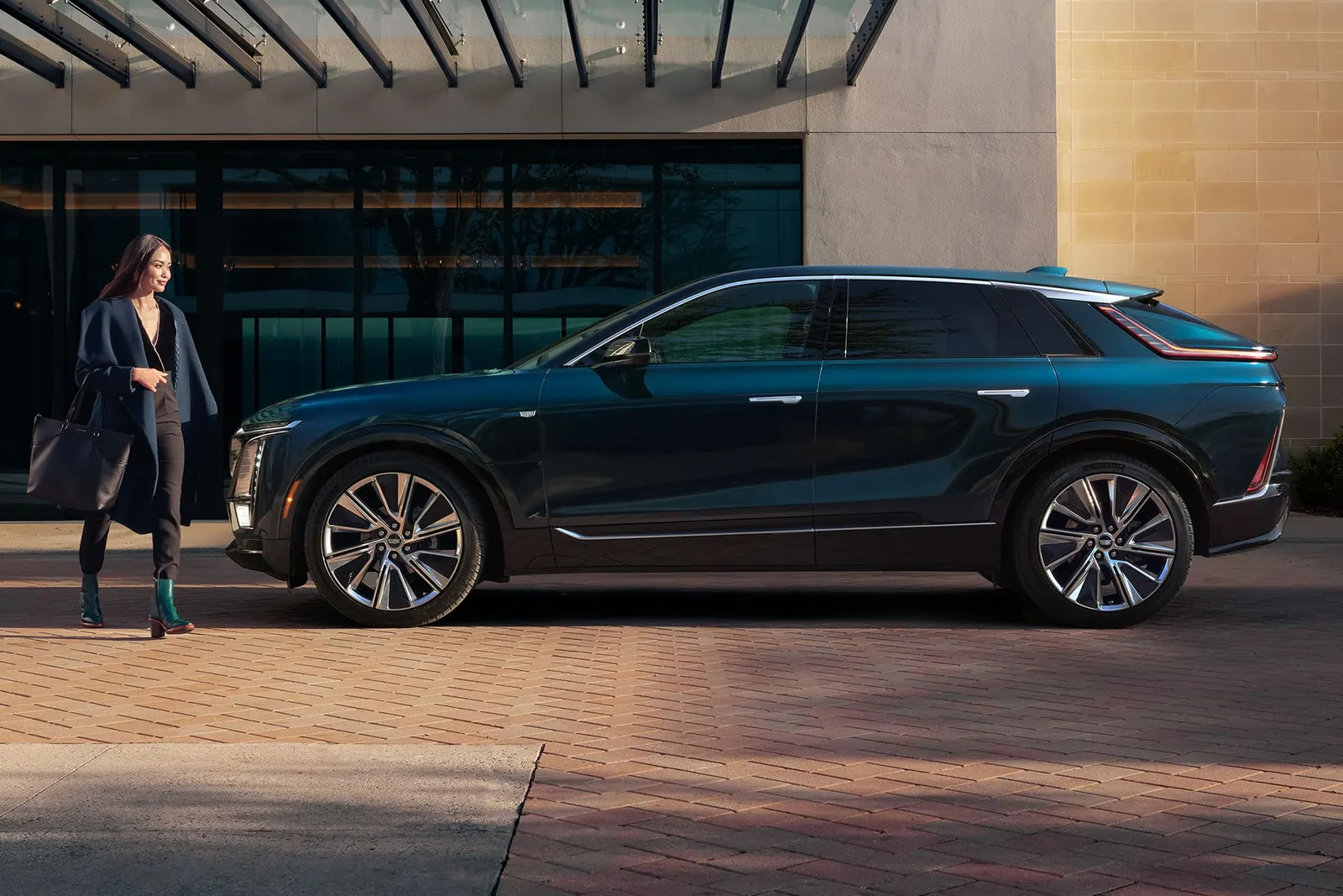 A dark 2027 Cadillac Lyriq parked in side profile outside a modern building while a woman walks toward it