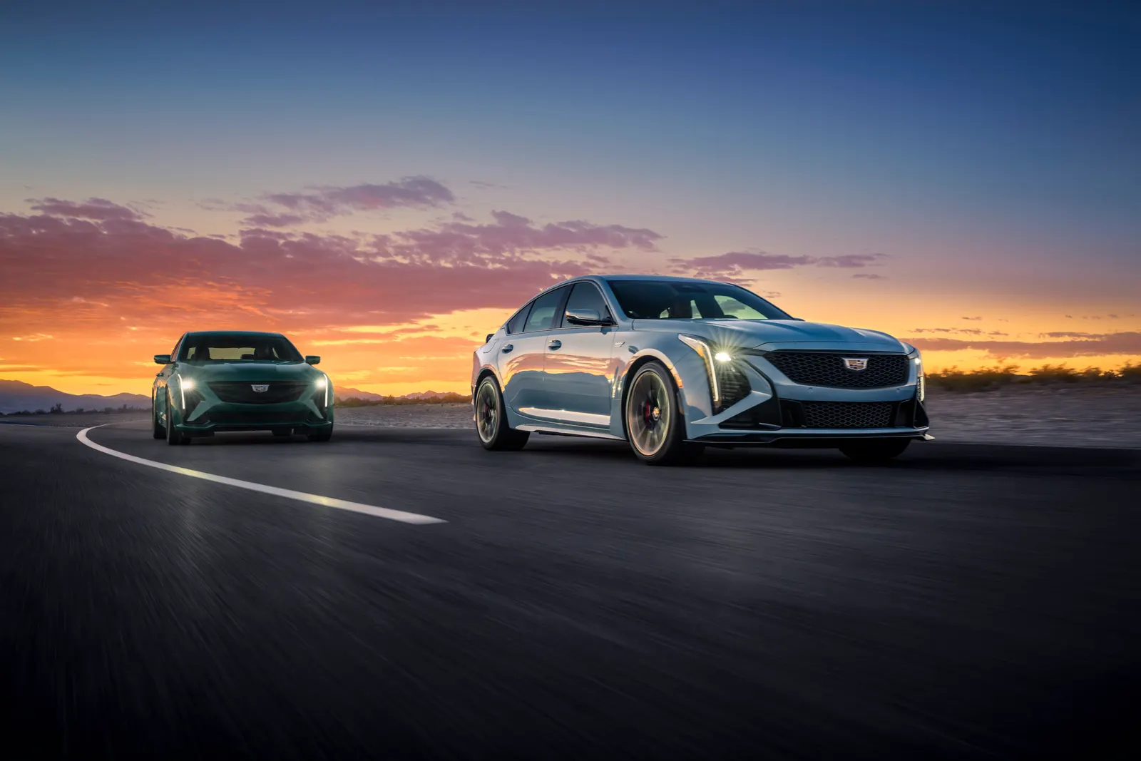 A silver and a green Cadillac CT5-V Blackwing driving on a desert road at dusk with their headlights on